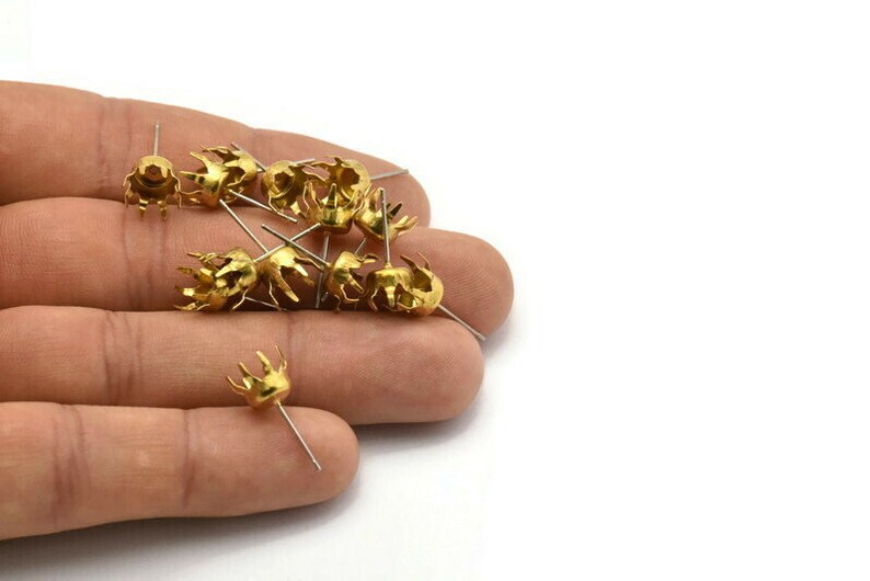 May include: A close-up of a hand holding a pile of gold-coloured metal crown-shaped settings with prongs. The settings are attached to small, thin metal pins.