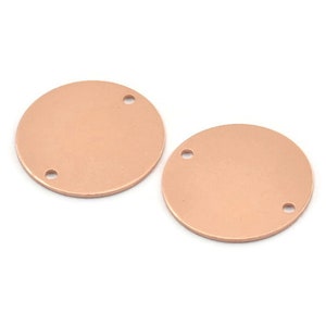Puede incluir: Dos discos redondos de metal color cobre, cada uno con dos agujeros, adecuados para la fabricación de joyas o manualidades. Los discos tienen un acabado liso y mate, sobre un fondo blanco.