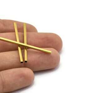 May include: Three gold-coloured metal tubes, approximately 3.8 cm long, are lying on a hand. The tubes are square in cross-section.