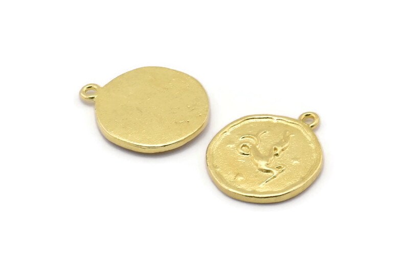 Op de afbeelding: Twee goudkleurige metalen bedels, een met een blanco ronde zijde en de andere met een reli&euml;f van een geit of ram. Beide bedels hebben een lus om aan een ketting of armband te bevestigen.