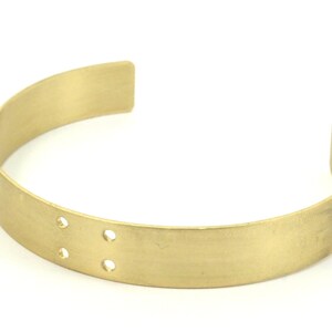 Op de afbeelding: Een eenvoudige gouden metalen armband met twee kleine gaatjes aan de onderkant. De armband is glad en heeft een licht afgeronde vorm.