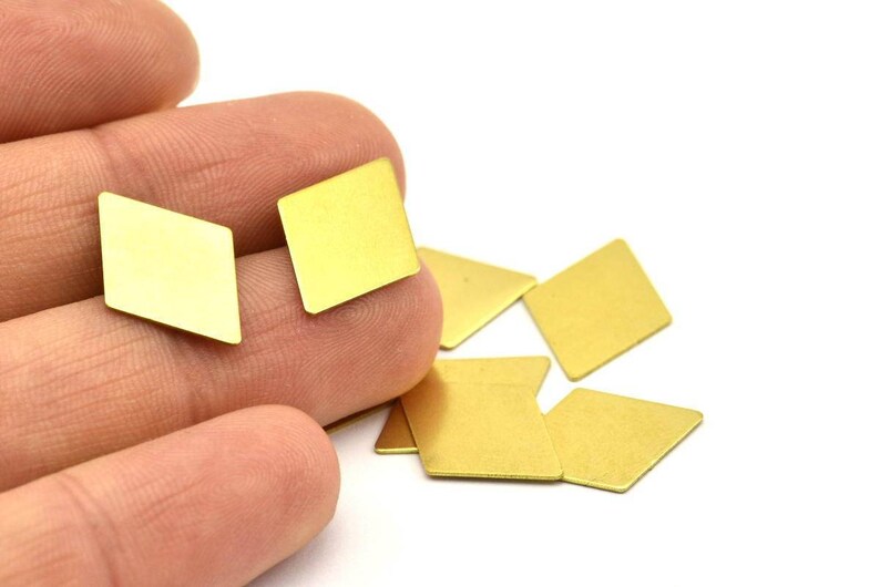 K&ouml;nnte beinhalten: Eine Gruppe kleiner, goldfarbener, rautenf&ouml;rmiger Metallst&uuml;cke. Die St&uuml;cke liegen auf einer wei&szlig;en Oberfl&auml;che verstreut und eine Hand h&auml;lt zwei der St&uuml;cke.