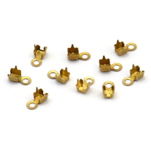 Puede incluir: Una colección de pequeños terminales de crimpado dorados para la fabricación de joyas. Cada terminal tiene forma rectangular con un lazo para sujetar a otros componentes. Los terminales están dispersos sobre un fondo blanco.