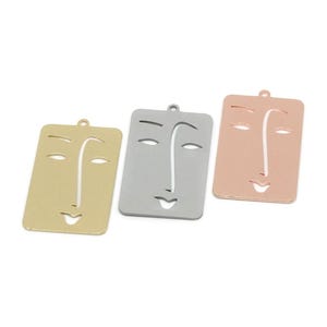 Puede incluir: Tres colgantes rectangulares de metal con un diseño de cara abstracta recortada. El conjunto incluye una pieza en tono dorado, una en tono plateado y una en tono oro rosa. Cada colgante tiene una pequeña anilla en la parte superior.
