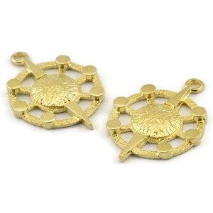 May include: Two gold-colored compass charms. Each charm features a central textured circle, a directional arrow, and a surrounding ring with evenly spaced circular accents. The charms have a loop at the top for attaching to jewelry.