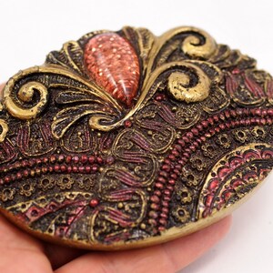May include: A gold-toned metal brooch with an intricate design featuring swirls, beads, and a red gem in the center.