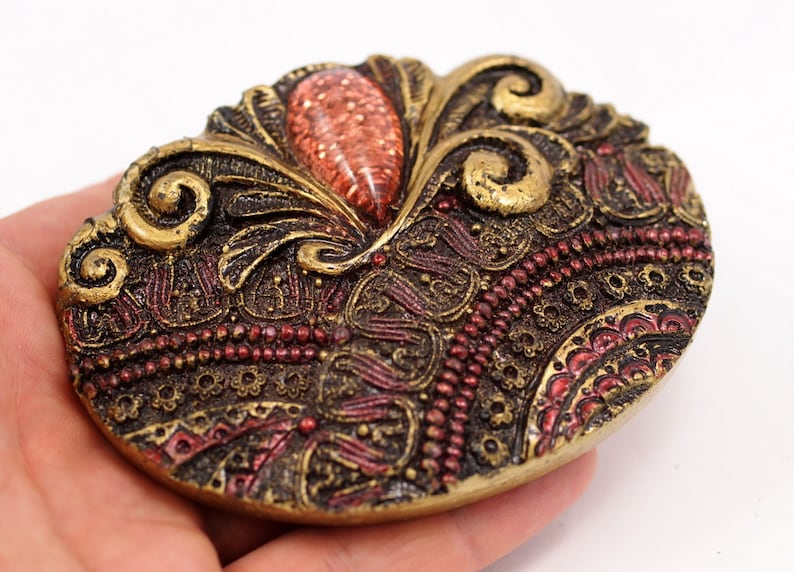 May include: A gold-toned metal brooch with an intricate design featuring swirls, beads, and a red gem in the center.