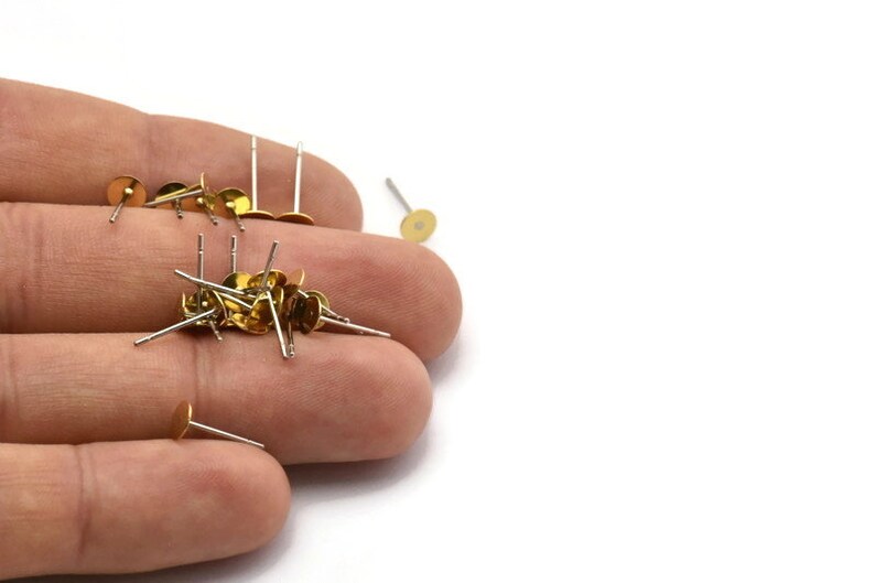 Puede incluir: Un mont&oacute;n de postes de pendientes de color dorado con tallos plateados. Los postes de pendientes est&aacute;n en una mano.