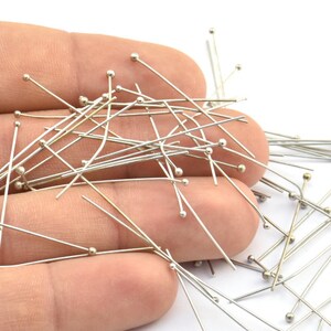 May include: A close-up of a hand holding a large quantity of silver-toned straight pins with round heads.