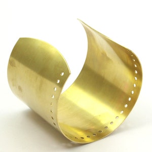 K&ouml;nnte beinhalten: Ein goldfarbenes Metall-Armband mit glatter, polierter Oberfl&auml;che. Das Armband hat eine Reihe kleiner L&ouml;cher entlang der Kante.