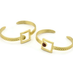 May include: Two gold bracelets with square-shaped accents. One bracelet has a red gemstone in the center of the square, while the other has a clear gemstone.
