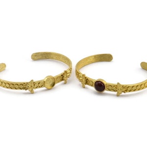 Könnte beinhalten: Zwei goldfarbene Armbänder mit einem Wabenmuster. Ein Armband hat eine runde, klare Steinfassung, das andere eine rote Steinfassung.