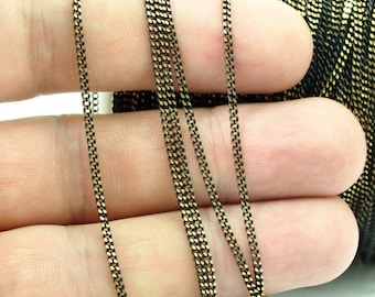 May include: Close-up of a black and gold chain. The chain is made of small, rectangular links, creating a textured surface. The chain is held in a hand, with a spool of the chain visible in the background.