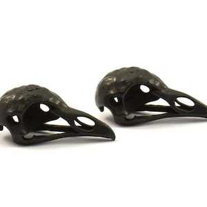以下が含まれることがあります： 黒い金属製の鳥の頭蓋骨のイヤリングが2つ。頭蓋骨は細部まで作り込まれており、質感のある仕上げになっています。