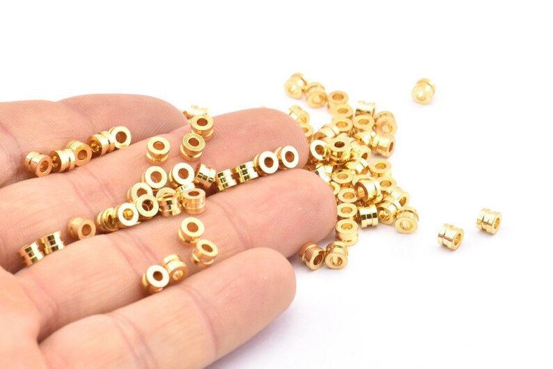 K&ouml;nnte beinhalten: Ein gro&szlig;er Haufen goldfarbener Metallperlen, von denen einige in einer Hand gehalten werden. Die Perlen sind rund und haben eine glatte Oberfl&auml;che.