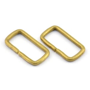 May include: Two gold-toned metal rectangular rings, also known as D-rings, are shown against a white background. These rings are commonly used for attaching straps, chains, or other hardware to bags, belts, or other leather goods.