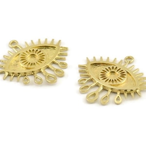 Puede incluir: Dos abalorios de metal dorado en forma de ojo con un diseño de sol en su interior. Los abalorios en forma de ojo tienen un borde elevado y una serie de pequeñas formas de lágrima alrededor del perímetro.