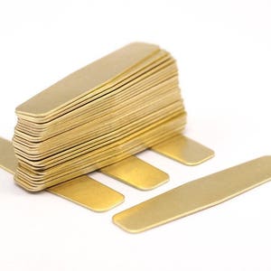 Puede incluir: Una pila de botones de camisa de metal de color dorado. Los botones de camisa son rectangulares y tienen bordes redondeados. Hay cuatro botones de camisa colocados planos sobre una superficie blanca.