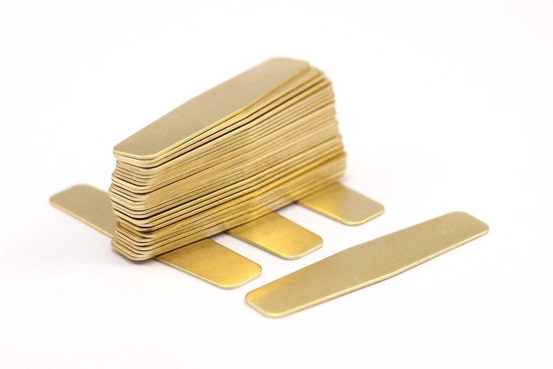 Puede incluir: Una pila de botones de camisa de metal de color dorado. Los botones de camisa son rectangulares y tienen bordes redondeados. Hay cuatro botones de camisa colocados planos sobre una superficie blanca.