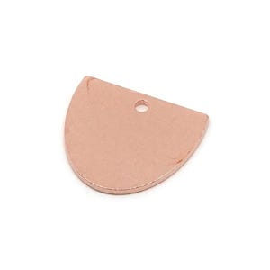 Puede incluir: Un colgante de color cobre, en forma de semicírculo, con un pequeño agujero en la parte superior para ensartar. El colgante tiene una superficie lisa y pulida y un borde inferior redondeado. Este componente de joyería es ideal para crear collares y pendientes.