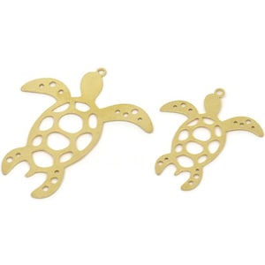 May include: Two gold-coloured metal turtle charms, one larger than the other, with a cutout design. The turtles are facing right and have a hole at the top for hanging.