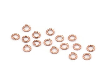 May include: A group of 16 rose gold jump rings, a type of jewelry finding, scattered on a white background.