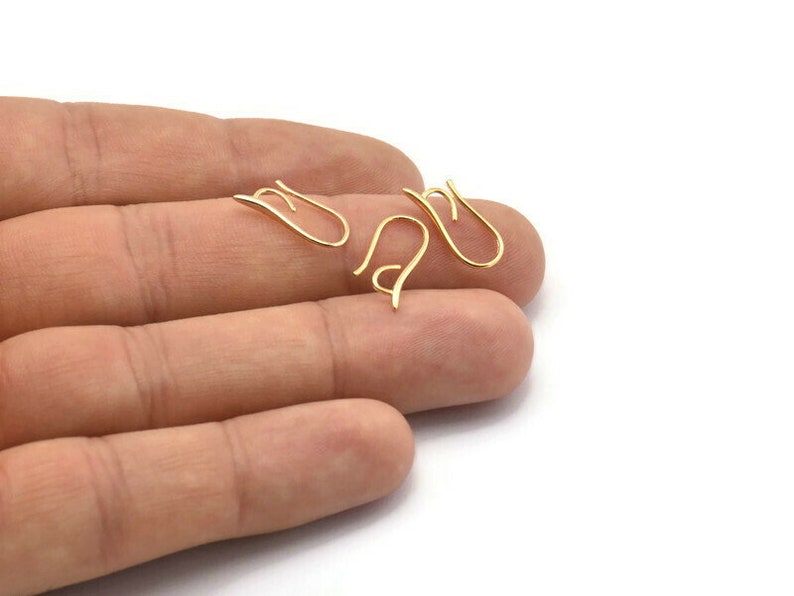 May include: Three gold-toned earring hooks, also known as ear wires, are shown on a hand. The hooks are made of metal and have a simple, elegant design.