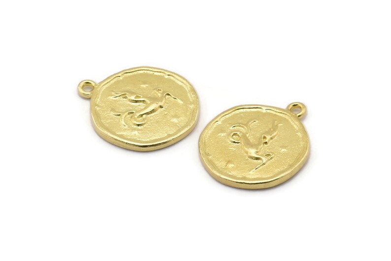 Op de afbeelding: Twee goudkleurige metalen bedels in de vorm van een cirkel met een reli&euml;fdesign van een gestileerde vogel. De bedels hebben een lus aan de bovenkant om aan sieraden te bevestigen.