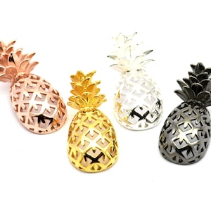 May include: Four pineapple-shaped metal charms in different colours: rose gold, gold, silver, and black. Each charm has a detailed pineapple design with a cutout pattern.