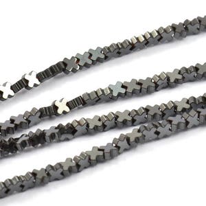Könnte beinhalten: Nahaufnahme von mehreren Strängen dunkelgrauer kreuzförmiger Perlen. Die Perlen sind aneinandergereiht und erzeugen ein strukturiertes, lineares Muster. Die Perlen haben einen metallischen Glanz und sind diagonal vor einem weißen Hintergrund angeordnet. Ideal für die Schmuckherstellung.