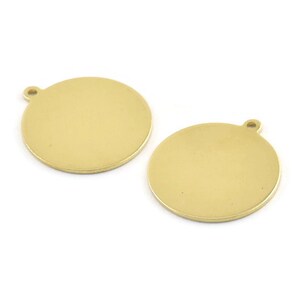 Puede incluir: Dos dijes redondos de metal de color dorado con un agujero en la parte superior para sujetar a un collar o pulsera.