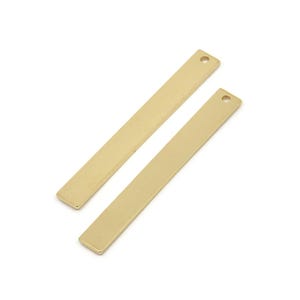 Puede incluir: Dos colgantes de barra de metal de color dorado con un agujero en la parte superior. Las barras son rectangulares y tienen un acabado liso y pulido.