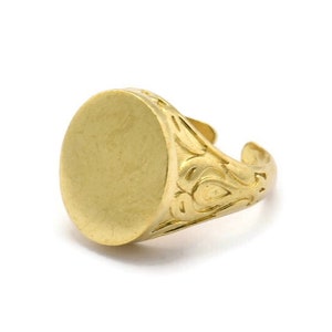 Puede incluir: Un anillo de sello dorado con una cara ovalada. El anillo presenta un diseño grabado y ornamentado en la banda. El anillo está sobre un fondo blanco.