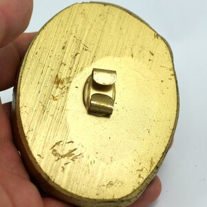 May include: A gold-colored, oval-shaped, metal pinback with a metal clasp on the back.