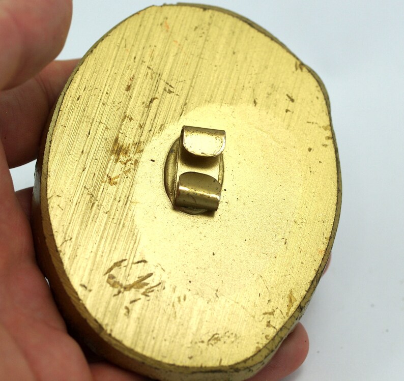 May include: A gold-colored, oval-shaped, metal pinback with a metal clasp on the back.