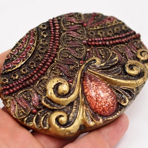 May include: A gold and red oval-shaped decorative piece with intricate details and a red gem-like center.