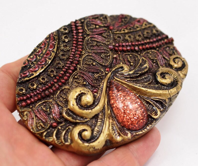 May include: A gold and red oval-shaped decorative piece with intricate details and a red gem-like center.