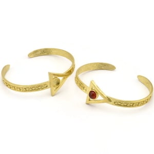 May include: Two gold bracelets with a triangle design and a red gemstone accent. The bracelets are made of metal and have a textured finish.