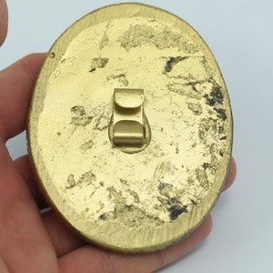 May include: Gold-colored metal pinback with a worn finish. The pinback has a metal clasp on the back.