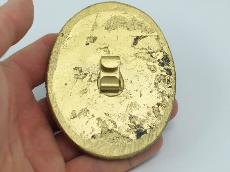 May include: Gold-colored metal pinback with a worn finish. The pinback has a metal clasp on the back.