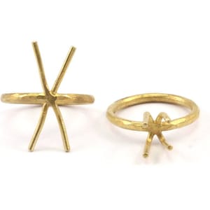 May include: Two gold-coloured ring settings with pronged designs. One ring has a six-prong setting, and the other has a four-prong setting. Both rings have a textured, hammered appearance against a white background.