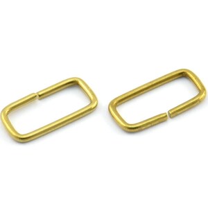 Puede incluir: Dos anillos rectangulares de metal de color dorado, también conocidos como anillos en D, se muestran sobre un fondo blanco. Los anillos están abiertos y listos para ser unidos a un proyecto.