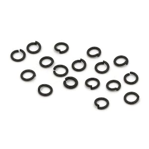 May include: A collection of small, black, open jump rings scattered on a white surface. These metal rings are commonly used in jewelry making and crafting. The rings are evenly spaced and appear to be made of a durable material.