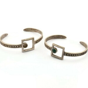 May include: Two silver cuff bracelets. Each bracelet features a square design with a small gemstone in the center. The bands are decorated with a series of small, square indentations. One bracelet has a brown gemstone, and the other has a green gemstone.