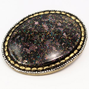 May include: A black oval brooch with a gold and silver beaded border. The center of the brooch is filled with iridescent glitter.