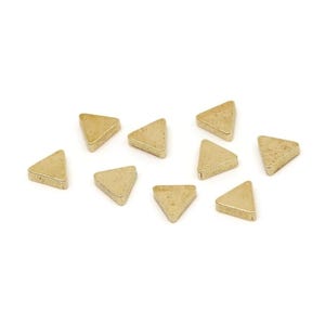May include: A collection of small, gold-colored triangle-shaped objects. Each triangle has a flat top and bottom with angled sides, and they are scattered on a white background. The objects appear to be made of metal.