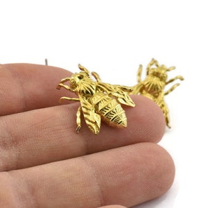 May include: Two gold-colored bee charms with detailed wings and bodies. The bees are on a hand, but the focus is on the charms.
