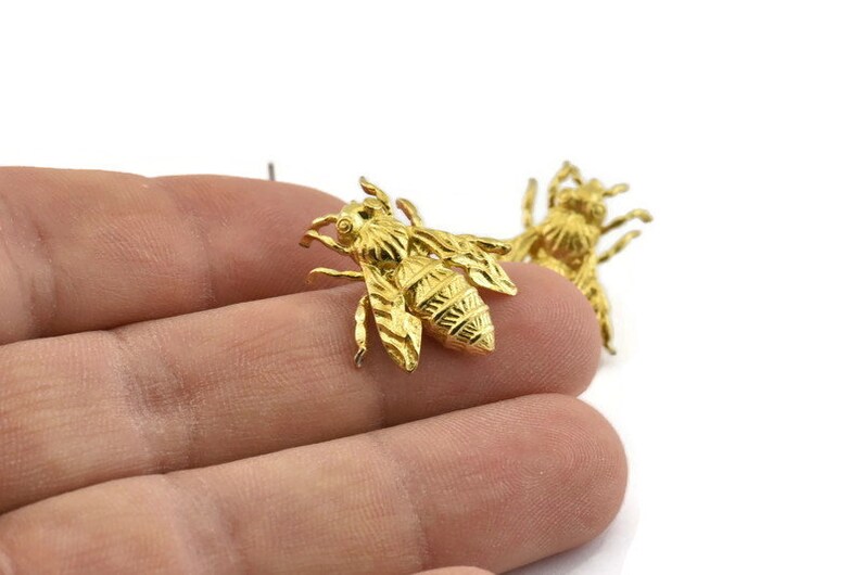 May include: Two gold-colored bee charms with detailed wings and bodies. The bees are on a hand, but the focus is on the charms.