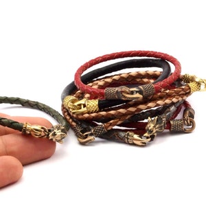K&ouml;nnte beinhalten: Ein Set aus f&uuml;nf Lederarmb&auml;ndern mit goldenen und messingfarbenen Akzenten. Die Armb&auml;nder sind geflochten und haben ein Wolfskopfdesign am Verschluss.