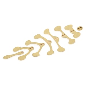 May include: A selection of gold-coloured, abstract bone-shaped charms with small, reddish-brown accents. The charms vary in size and shape, arranged in a pattern against a white background. These charms could be used for jewellery making or other craft projects.